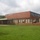 The image shows a brick building identified as 'N.G. Armory' (National Guard Armory), a mid-20th century institutional structure with a distinctive tall chimney stack on the right side. The building features a low-profile design with clerestory windows on the upper section and window-mounted air conditioning units. The facility is surrounded by a well-maintained green lawn under a partly cloudy sky.