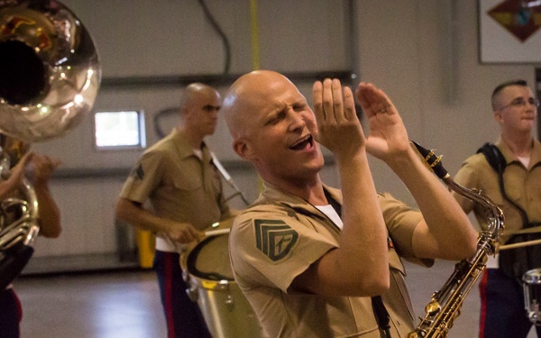 A group of U.S. Marine Corps band members are rehearsing or performing inside what appears to be a gymnasium or indoor drill facility. The central figure, a bald Marine holding a saxophone, is singing or calling out with his hands raised expressively. Other band members are visible playing a tuba, snare drum, and tenor drum in the background.