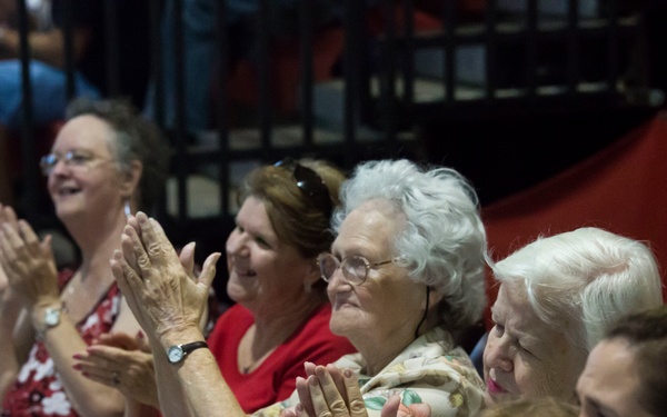 A group of women of various ages, including elderly women and younger adults, are seated in what appears to be an auditorium or event venue. They are enthusiastically clapping and smiling, suggesting they are watching a performance or presentation. Several are wearing red, white, and blue clothing, indicating a patriotic-themed event.