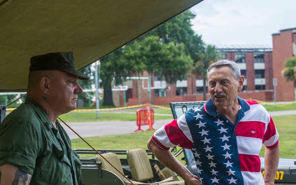 Two men are conversing at an outdoor military history display or exhibit, set up under a canvas canopy on a grassy area. One man is dressed in green military-style fatigues with a military cap and visible tattoos, while the other wears an American flag-patterned polo shirt. A vintage military Jeep (likely a WWII-era Willys MB) is parked in the background, and a table with military artifacts is visible in the foreground.