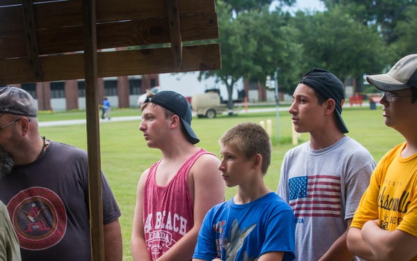 A group of young males and an adult man are gathered under a covered outdoor display booth, appearing to listen to a presenter in military attire. The setting appears to be an outdoor military or historical exhibition event on a grassy field, with institutional buildings and an inflatable bounce structure visible in the background. A table in the foreground holds military equipment, gear, and artifacts for display.