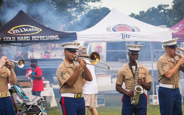 Four U.S. Marine Corps musicians in dress uniform are performing at an outdoor public event, playing brass and woodwind instruments on a grass field. The setting appears to be a festival or community gathering with vendor tents, spectators, and barbecue smoke visible in the background. Brand tents for Mike's Hard Lemonade, Miller Lite, and Yuengling are prominently displayed behind the performers.