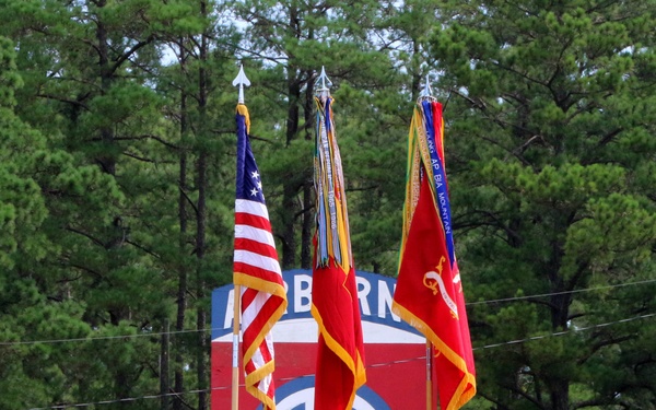 A military color guard ceremony featuring five soldiers in U.S. Army OCP (Operational Camouflage Pattern) uniforms and maroon berets, standing at attention on a grass field. Three soldiers hold ceremonial flags including the U.S. national flag and red unit guidons, while two armed soldiers stand as rifle guards on the flanks. The backdrop features a large red sign displaying the 82nd Airborne Division insignia with the double 'AA' (All American) patch.
