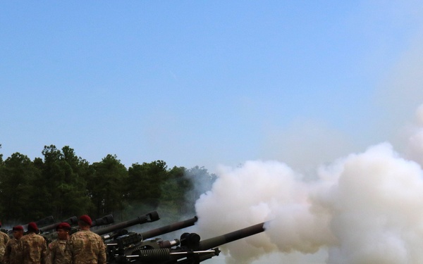 A ceremonial artillery salute is being conducted by soldiers of the 82nd Airborne Division, identifiable by the red and white 'AA' (All American) Airborne sign visible in the background. Multiple howitzers are lined up on a grass field, with at least one cannon visibly firing, producing a large white smoke cloud. The soldiers, wearing red berets and camouflage uniforms, are standing at attention beside the artillery pieces.