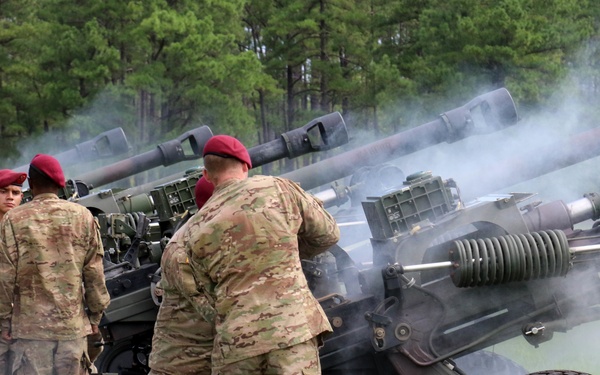 A group of military soldiers wearing camouflage uniforms and distinctive red berets are operating multiple artillery pieces on a grassy field. Smoke is visible near one of the cannons, indicating a recent or ongoing firing sequence. The setting appears to be a ceremonial or training artillery salute exercise, likely at a U.S. Army installation.