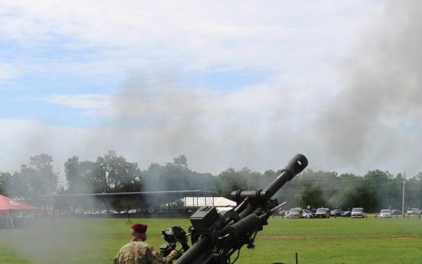 A military soldier in camouflage uniform and red beret operates an artillery howitzer on a grassy field during what appears to be a ceremonial or demonstration event. The cannon has recently been fired, as evidenced by the smoke rising from the barrel and dispersing into the overcast sky. A crowd of spectators is visible in the background under a red tent, with parked vehicles lining the perimeter of the field.