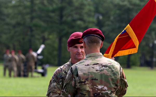 A military ceremony is taking place on a grassy field, featuring soldiers wearing maroon berets and camouflage uniforms. Two soldiers in the foreground appear to be engaged in a formal exchange, with one holding a red ceremonial flag with gold fringe. Additional soldiers and what appears to be an artillery piece are visible in the background.