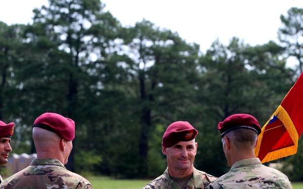 Four U.S. Army soldiers wearing maroon berets and OCP (Operational Camouflage Pattern) uniforms are gathered on a grassy field during what appears to be a military change of command or unit ceremony. A guidon or unit colors flag with red, yellow, and gold colors adorned with multiple streamer ribbons is visible on the right side. The setting and attire suggest this is an 82nd Airborne Division formal military ceremony.