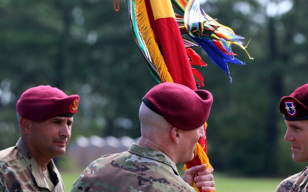 Three U.S. Army soldiers wearing maroon berets and OCP (Operational Camouflage Pattern) uniforms are participating in a formal military change of command ceremony. A guidon (unit flag) with streamers is being passed between two soldiers, indicating a transfer of command authority. The setting appears to be an outdoor parade field with troops formed in the background.