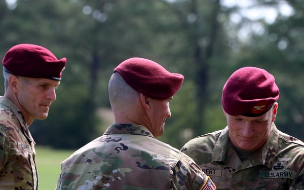 Three U.S. Army soldiers wearing maroon berets and OCP (Operational Camouflage Pattern) uniforms are gathered outdoors in what appears to be a military ceremony or award presentation. Two soldiers face each other in the foreground while a third observes from the left. The maroon berets and 82nd Airborne Division patches indicate these are paratroopers, likely at Fort Bragg (now Fort Liberty), North Carolina.