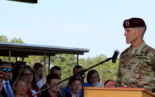 A U.S. Army officer wearing a maroon beret and camouflage uniform with an 82nd Airborne Division patch speaks at a wooden podium during what appears to be a formal military ceremony. An audience of military personnel and civilians, including families, is seated behind him under a covered pavilion. The outdoor setting features flowering rose bushes in the foreground.