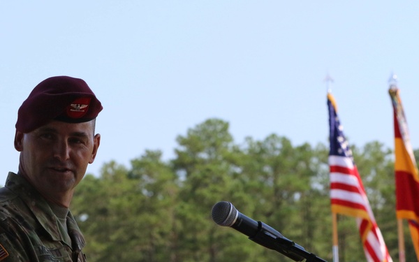 A U.S. Army officer wearing a maroon beret and camouflage uniform stands at a podium with a microphone, appearing to deliver a speech at a formal military ceremony. The background features multiple flags including the U.S. flag and unit colors, along with a sign displaying the '82nd Airborne Division' double-A insignia. The outdoor setting with pine trees and clear sky suggests a daytime ceremonial event at Fort Bragg (now Fort Liberty), North Carolina.
