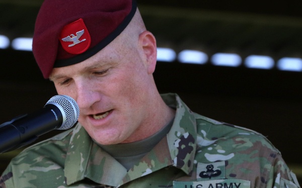 A U.S. Army Colonel named Hilbert is speaking at a podium microphone during what appears to be a formal military ceremony. He is wearing an OCP (Operational Camouflage Pattern) uniform with a maroon beret, indicating airborne affiliation, and displays the 82nd Airborne Division patch on his right shoulder. Red flowers are visible in the foreground, suggesting an outdoor ceremonial setting.