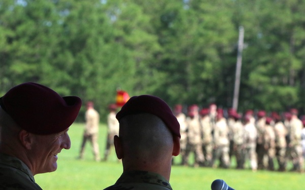 Two senior military personnel wearing maroon berets stand at a podium with a microphone, observing a formation of soldiers on a grassy field. The soldiers in the background are dressed in tan/multicam uniforms and also wear maroon berets, suggesting a airborne or paratrooper unit ceremony. The setting appears to be an outdoor military parade or change of command event.