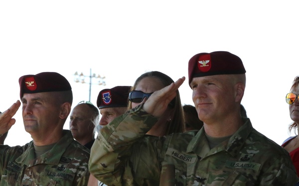 Two U.S. Army soldiers wearing maroon berets and OCP (Operational Camouflage Pattern) uniforms are rendering a salute at what appears to be a military ceremony. The name tape 'HILBERT' is visible on the right soldier's uniform. Civilians, including women in dress attire, stand alongside and behind the soldiers, suggesting a formal change of command or unit ceremony.
