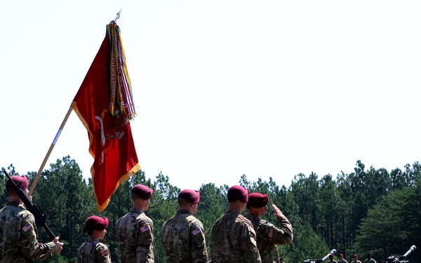 A military ceremony is taking place on an open grassy field, with soldiers in camouflage uniforms and distinctive maroon/crimson berets marching in formation. A color guard carries a red unit guidon/flag with gold fringe and campaign streamers. Artillery pieces and additional troops are visible in the background, suggesting a formal change of command or unit activation ceremony.