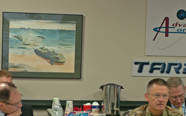 A formal meeting is taking place at TARDEC (Tank Automotive Research, Development and Engineering Center) in a conference room setting. Visible name placards identify attendees including Dr. Paul Rogers and Daniel Allyn, with military personnel in Army combat uniforms seated alongside civilians in business attire. The room displays 'Advanced Concepts' and 'TARDEC' signage along with military vehicle artwork on the wall.