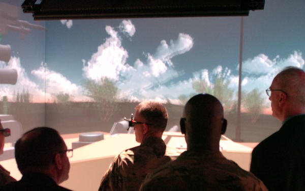 A group of individuals, including several in military camouflage uniforms and at least two in civilian attire, are viewing a large-scale immersive simulation environment. The simulation displays a desert-like outdoor scene with blue sky, clouds, and vegetation projected on surrounding screens. Some participants appear to be wearing 3D glasses, suggesting an interactive or stereoscopic display system.