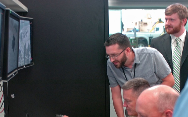A group of individuals, including at least two military personnel in camouflage uniforms and several civilians, are gathered around a workstation with multiple monitors displaying aerial or surveillance imagery. One military person is operating a joystick controller while others observe. The setting appears to be a control room or demonstration facility with cinder block walls.