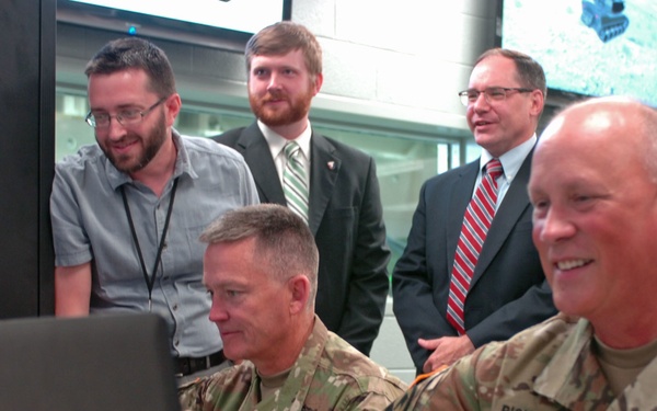 A group of individuals, including two U.S. Army soldiers in camouflage uniforms and three civilians in business attire, are gathered around laptop computers in what appears to be a military operations or technology center. The soldiers and civilians appear engaged and smiling while reviewing content on the screens. A name tag reading 'RICHARDSON' and 'U.S. ARMY' is visible on the soldier in the foreground.