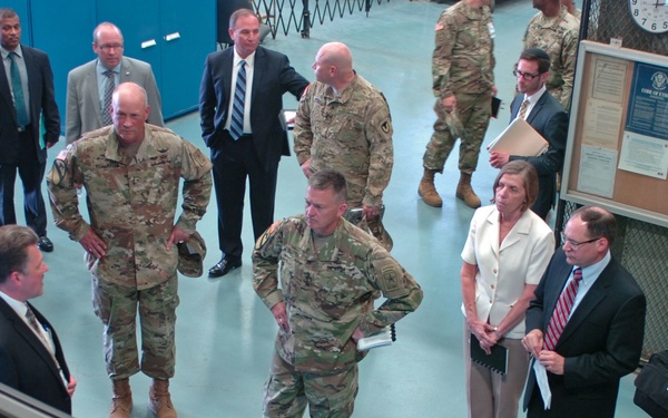 A formal briefing or tour is taking place inside what appears to be a military facility or operations center. A mixed group of military personnel in camouflage uniforms and civilians in business attire are gathered around a presenter near computer monitors. The setting suggests an official visit, inspection, or command briefing.