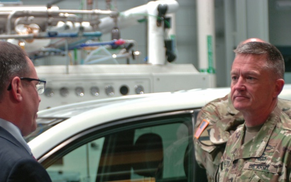 A U.S. Army soldier in MultiCam/OCP camouflage uniform is engaged in conversation with a civilian man in a dark suit inside what appears to be a technology or research facility. A vehicle with 'FUEL' visible on its door panel is positioned between them, suggesting this may be a demonstration or briefing related to fuel cell or alternative energy vehicle technology. The setting includes industrial piping and equipment visible in the background.