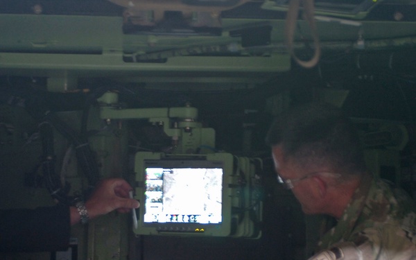 Two individuals are seated inside what appears to be a military vehicle, examining a mounted display screen. One person is wearing a civilian business suit while the other is in military camouflage (MultiCam/OCP pattern) uniform. They appear to be reviewing tactical or surveillance imagery on the vehicle-mounted monitor.