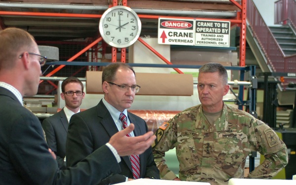 A group of military and civilian personnel are gathered inside an industrial facility for what appears to be a briefing or tour. A civilian in a dark suit is presenting or explaining something to attendees including at least one uniformed U.S. Army officer and other suited individuals. White packaged boxes with labeled identifiers are visible on a table in the foreground.