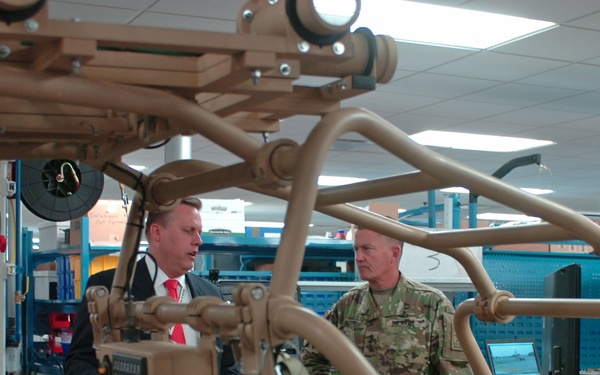 Two individuals — one in a business suit with a red tie and one in military camouflage uniform — are engaged in a discussion inside what appears to be a military equipment facility or laboratory. They are standing near tan/desert-colored military vehicles or vehicle frames, likely light tactical vehicles or similar equipment. The setting suggests a briefing or inspection tour of military hardware.