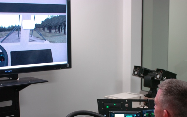 A military personnel in camouflage uniform is operating what appears to be a vehicle driving simulator. The setup includes a steering wheel, pedals, multiple control panels with displays, and a large Sony monitor showing a first-person driving perspective through a tree-lined road. The environment appears to be an indoor simulation or testing facility.