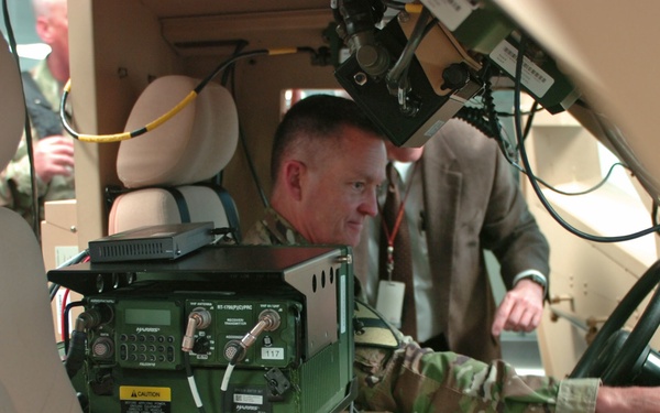 The image shows the interior of a military vehicle, likely a HMMWV (Humvee), equipped with multiple Harris brand military communications and radio systems. A uniformed military personnel is seated inside examining the equipment, while a civilian in a suit with a lanyard badge observes from behind. The setting appears to be a demonstration or exhibition of military communications technology.