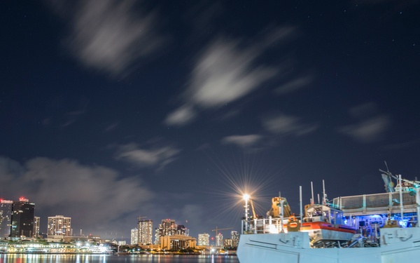 A nighttime long-exposure photograph of a large white vessel named 'STRATTON' docked at a pier, with a illuminated city skyline reflected in calm harbor waters. The image captures streaking clouds in a dark blue sky, suggesting an extended shutter exposure. The scene is set in what appears to be a major port city with high-rise buildings visible in the background.