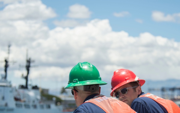 Three personnel wearing orange life vests and hard hats (two green, one red) are leaning against a railing and observing a harbor. A U.S. Coast Guard vessel is visible in the background, docked at what appears to be a Coast Guard station or port facility. The scene takes place under partly cloudy skies with calm blue water visible.
