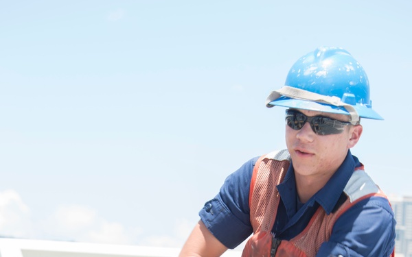 A young male worker is kneeling on a vessel deck, gripping and pulling a thick white mooring rope with both hands. He is wearing standard maritime safety gear including a blue hard hat, orange life vest, blue coveralls, and dark safety glasses. The setting appears to be the deck of a ship or large vessel in bright daylight conditions.