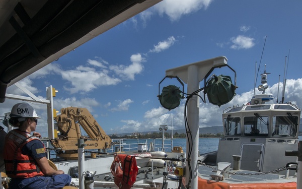 The image shows a U.S. Coast Guard vessel deck with personnel in safety gear and hard hats. Multiple boats are visible including a gray patrol boat and an orange rescue craft, set against a harbor backdrop with blue skies and partial cloud cover. Equipment, life vests, and rigging are prominently displayed across the deck.