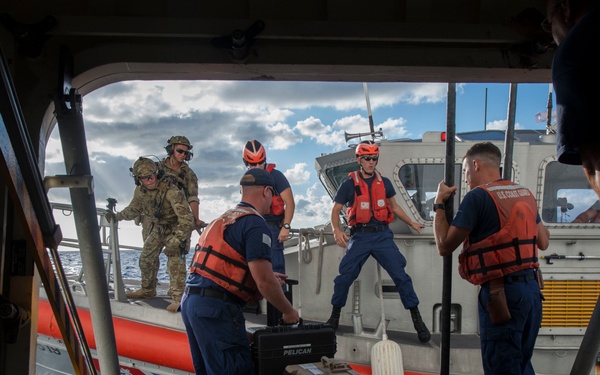 The image shows a U.S. Coast Guard operation involving personnel transferring equipment between a larger vessel and a smaller rigid-hull inflatable boat (RHIB). Multiple personnel wearing orange life vests and blue uniforms are handling Pelican cases and other gear during what appears to be a maritime logistics or boarding operation. One individual in military tactical gear and helmet is also present on the smaller vessel.