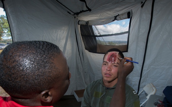 A person in a red shirt is applying simulated wound makeup/prosthetics to a man wearing an olive green military flight suit inside a white tent. The realistic-looking fake head wound on the subject's forehead is being detailed with a small brush. A table nearby holds makeup supplies, fake blood, and prosthetic materials, indicating this is a medical training or disaster simulation exercise.
