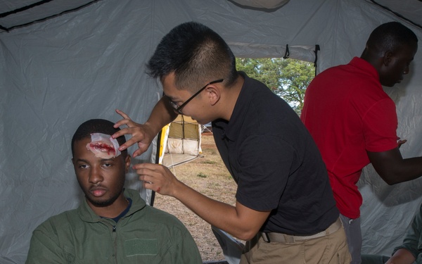 The image shows what appears to be a military training exercise involving simulated casualty makeup application inside a field tent. A person in a black shirt and glasses is applying simulated wound makeup to the forehead of a seated individual in an olive green military jumpsuit. In the background, a second person in a red shirt is applying similar moulage makeup to another seated individual in military attire.