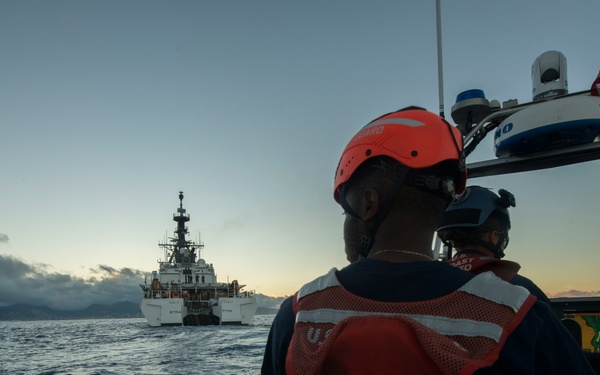 A small U.S. Coast Guard boat crew approaches a larger Coast Guard cutter vessel on open ocean waters at dusk or dawn. Personnel aboard the small boat are wearing orange life vests and helmets, suggesting an active operational or training exercise. A mountainous coastline is visible in the background under a partly cloudy sky.