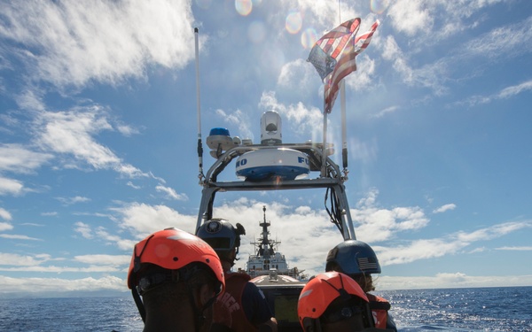 A small rigid inflatable boat (RIB) is underway at speed on open ocean, carrying several personnel wearing orange life vests and orange helmets. The crew appears to be approaching a larger Coast Guard cutter visible in the background. An American flag is flying from the boat's mast, and the scene suggests a maritime law enforcement or rescue operation.