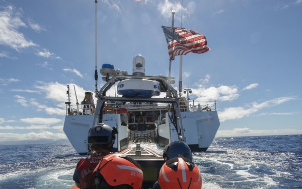 A U.S. Coast Guard small inflatable rescue boat with multiple crew members in orange life vests and helmets approaches a larger Coast Guard vessel flying the American flag on open ocean waters. The crew members are wearing clearly marked 'U.S. Coast Guard' gear and appear to be conducting a training exercise or operational approach maneuver. The scene takes place under partly cloudy skies with choppy blue ocean waters.