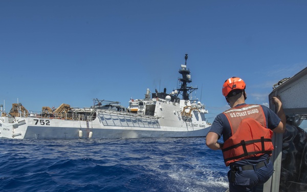 A U.S. Coast Guard personnel stands on a small response boat observing a larger Coast Guard cutter numbered 752 in open blue ocean waters. The crew member is wearing standard Coast Guard operational gear including an orange life vest and helmet. The scene depicts an active maritime operation with both vessels underway.