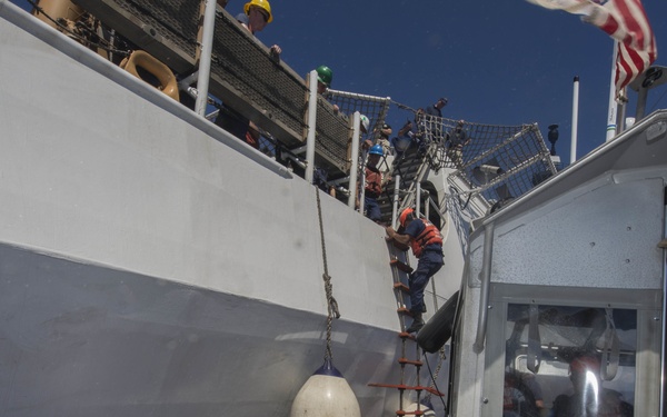 A maritime boarding operation is underway, with personnel climbing a rope/Jacob's ladder from a smaller vessel onto the side of a larger gray ship. The smaller craft, likely a rigid-hull inflatable boat (RHIB) or patrol vessel, is positioned alongside the larger ship which has partial hull markings visible reading 'PROJ' and numbers '26', '5', '4'. An American flag is visible in the upper right, suggesting a U.S. government or military/Coast Guard operation.
