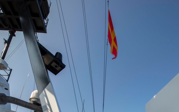 Two naval personnel wearing green helmets and orange life vests are on the deck of a military vessel at sea. One crew member is hoisting a signal flag — red and yellow, consistent with the NATO/International Code of Signals 'N' (November) pennant or a similar naval signal flag — while the other observes. The scene takes place on a clear day with calm blue ocean visible in the background.