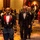 A formal military ceremonial event taking place in a large ballroom or banquet hall. Three prominent figures are walking in procession down a carpeted aisle, with two in military dress uniforms and one in civilian formal attire (tuxedo). An American flag and what appears to be a state flag are visible in the background, along with a color guard and assembled guests.
