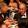 Two men are seated at a formal dinner table engaged in close conversation. One man is wearing a U.S. Army dress uniform with multiple service ribbons and medals, while the other is dressed in a black tuxedo with a bow tie. The setting appears to be a formal military banquet or gala event with multiple guests visible in the background.