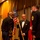 A formal military ceremony taking place on a stage, likely a dining-in or military ball event. A civilian in a tuxedo is holding a military guidon/flag staff while interacting with two U.S. Army soldiers in dress uniforms. The setting features gold curtains, multiple flags including the U.S. flag and what appears to be a general officer's flag.