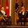 Two men are seated on a stage in leather armchairs during what appears to be a formal military ceremony or gala. One man is wearing a U.S. Army dress uniform with medals and decorations, while the other is in a black tuxedo and holding a microphone, suggesting an interview or moderated discussion format. Multiple military flags and unit colors are displayed in the background.