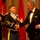 A formal military ceremony or gala event where a uniformed military officer is shaking hands with a civilian man in a tuxedo while holding a microphone. A second uniformed officer stands in the background applauding. The setting appears to be an official banquet or awards ceremony with multiple military flags displayed.