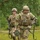 Two U.S. Army soldiers in Operational Camouflage Pattern (OCP) uniforms are running across a grassy field during what appears to be a training exercise. The soldier in front is identified by the name tape 'GUERRERO' and carries a rifle, while the soldier behind, identified as 'THOMAS', carries both a rifle and a green military ammunition can. Both wear combat helmets and protective eyewear.